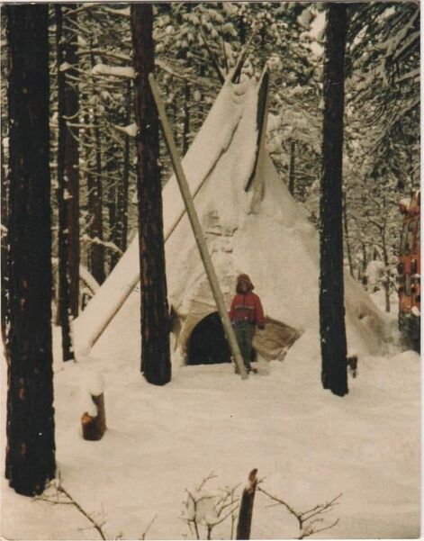 Picture is faded and early 80's in appearance. It is of a child standing in front of a tipi in the middle of the pine forest, all covered in snow. There is the tail end of a bus to the right of the picture. Three trees in the foreground, and a fallen tree a couple steps before the child is leaned against another tree. Child is in a faded red coat zipped up the front with a white strip over a blue strip across the front of it and a pair of jeans. All one can see is a paler line of face from under the hood. Time of year appears undetermined but the snow appears either fresh and a couple inches. The door to the tipi is open and it is dark inside. 