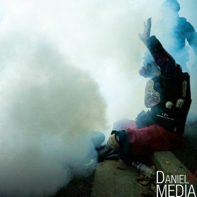 Picture of human wearing motorcycle armor over a red dress. Human has gas mask on and their hands raised as they sit on a curb. They are sitting in a thick cloud of tear gas with an officer standing behind them.